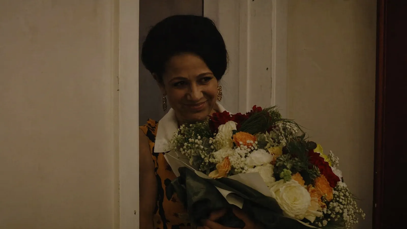 Una mujer sonriente de pie junto a una puerta, con un gran ramo de flores en los brazos