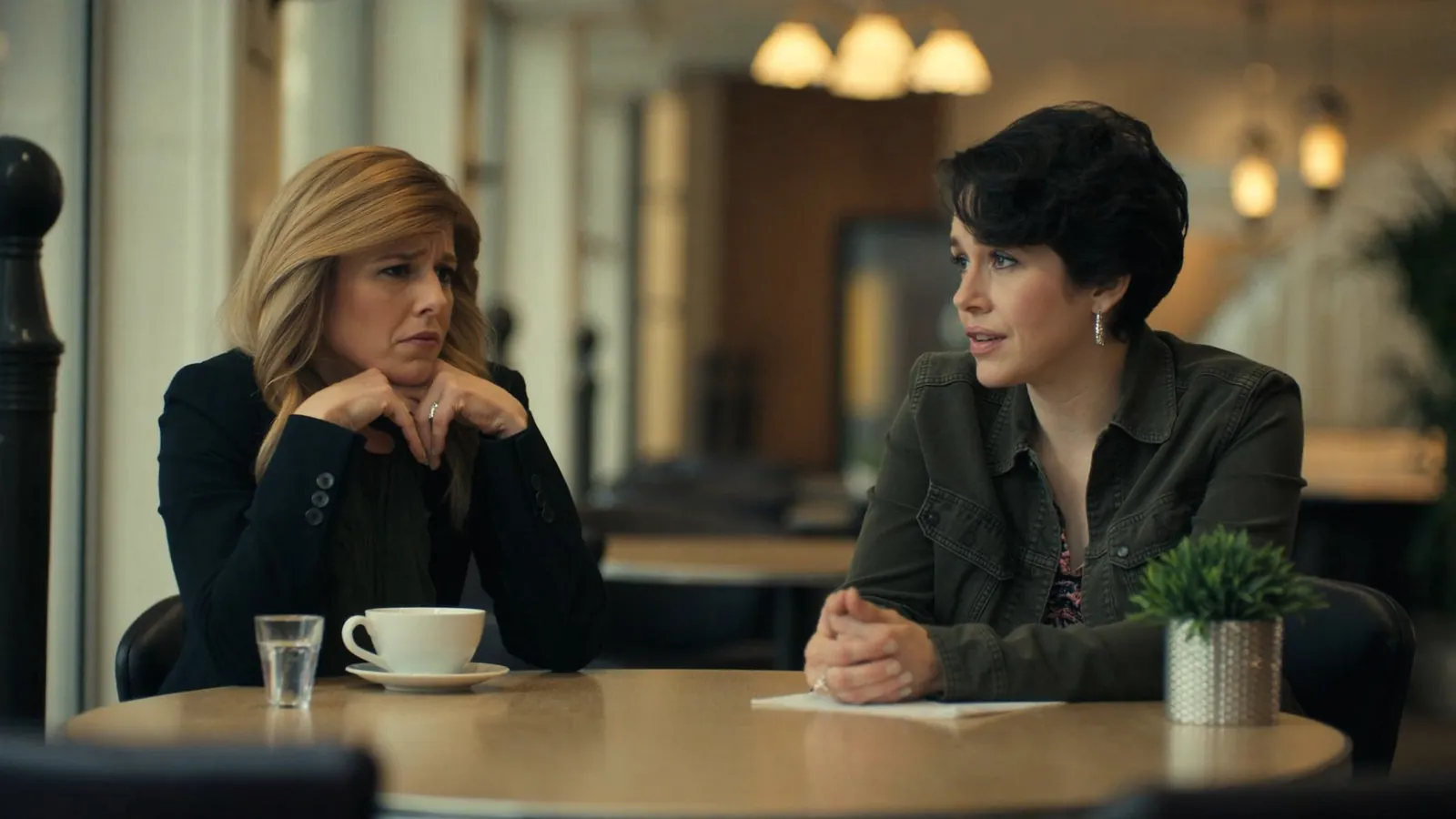 Dos mujeres sentadas frente a frente en la mesa de una cafetería, una hablando y la otra escuchando con gesto serio