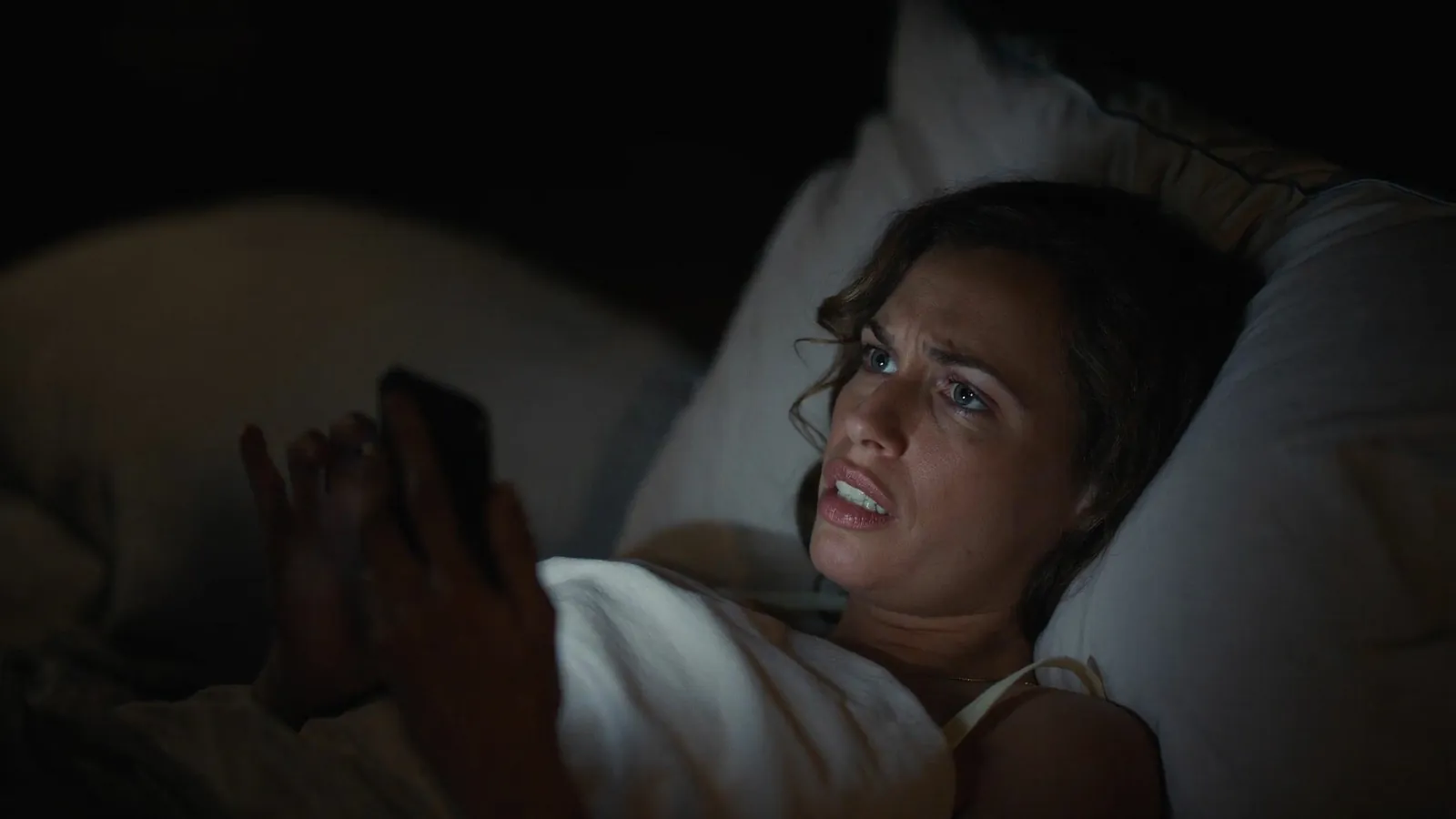 Una mujer tumbada en la cama de noche, con la cara iluminada por el móvil y gesto de incredulidad
