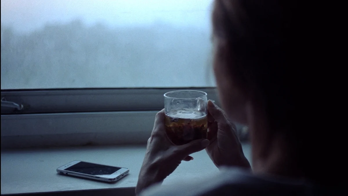 Junto a una ventana con niebla, una mujer sostiene un vaso entre las manos mientras el móvil descansa en el alféizar, sin tocarlo