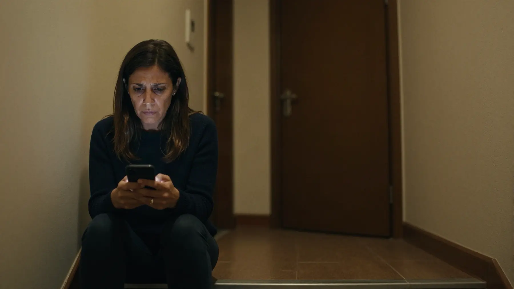 Una mujer sentada sola en la escalera de un portal por la noche, con el móvil entre las manos y gesto confuso