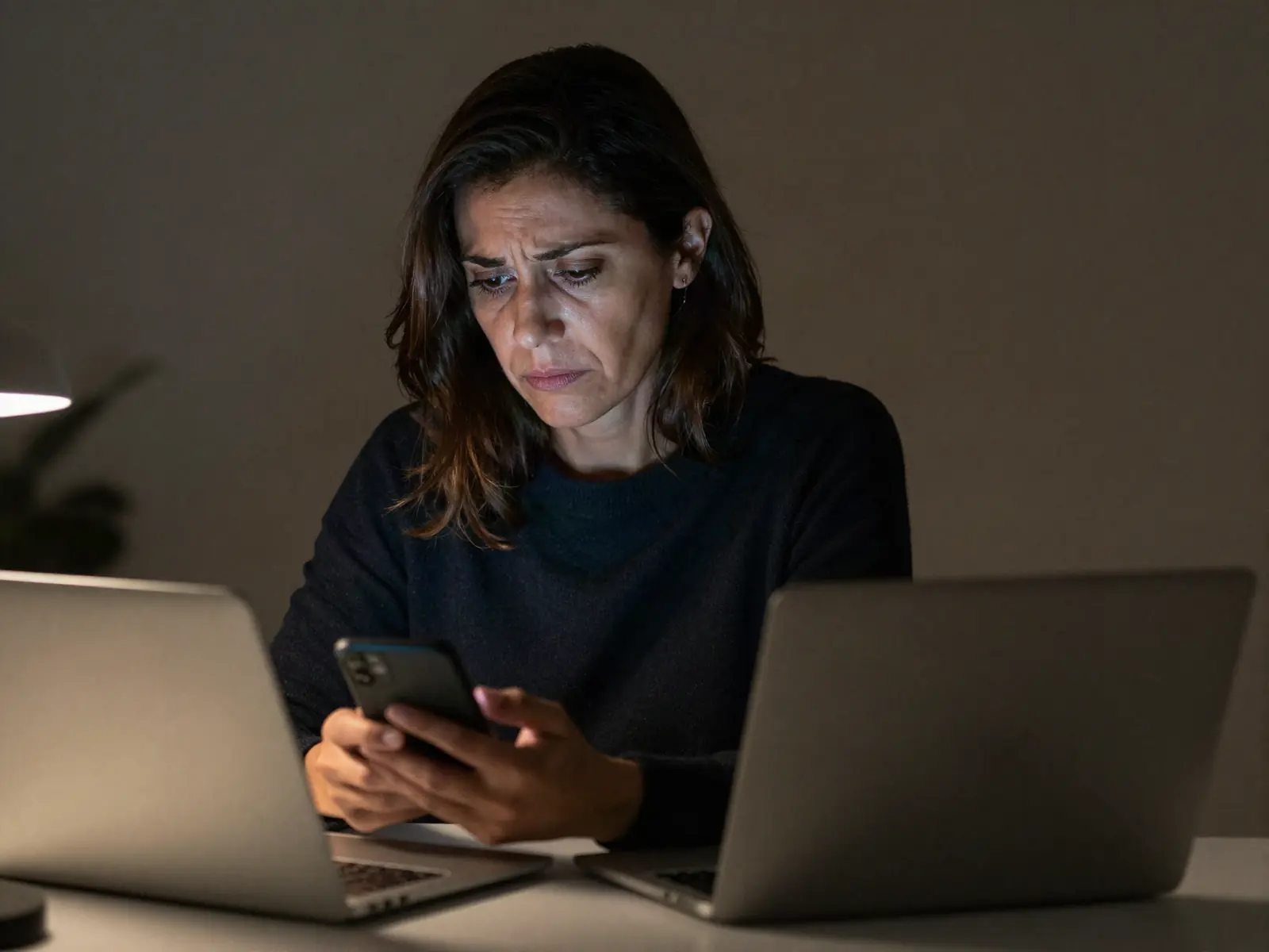 Una mujer ante un portátil cerrado, releyendo mensajes en el móvil por la noche, con gesto cansado y confuso