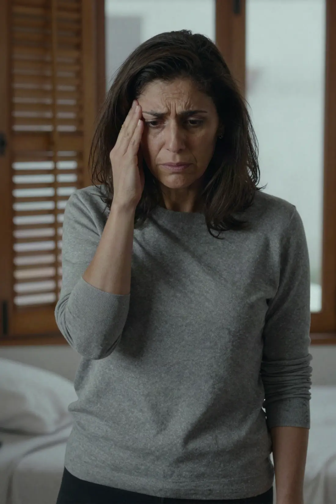 Una mujer morena de unos cuarenta años, de pie junto a una ventana con contraventanas de madera, con la mano en la sien y gesto confuso